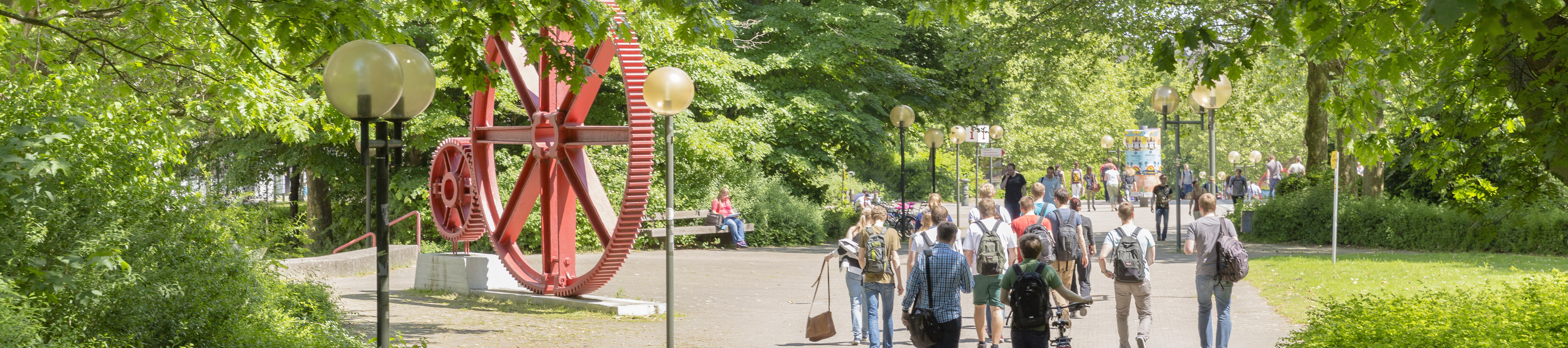 Studenten im Sommer auf dem Campus Nord, rote Zahnräder im Hintergrund